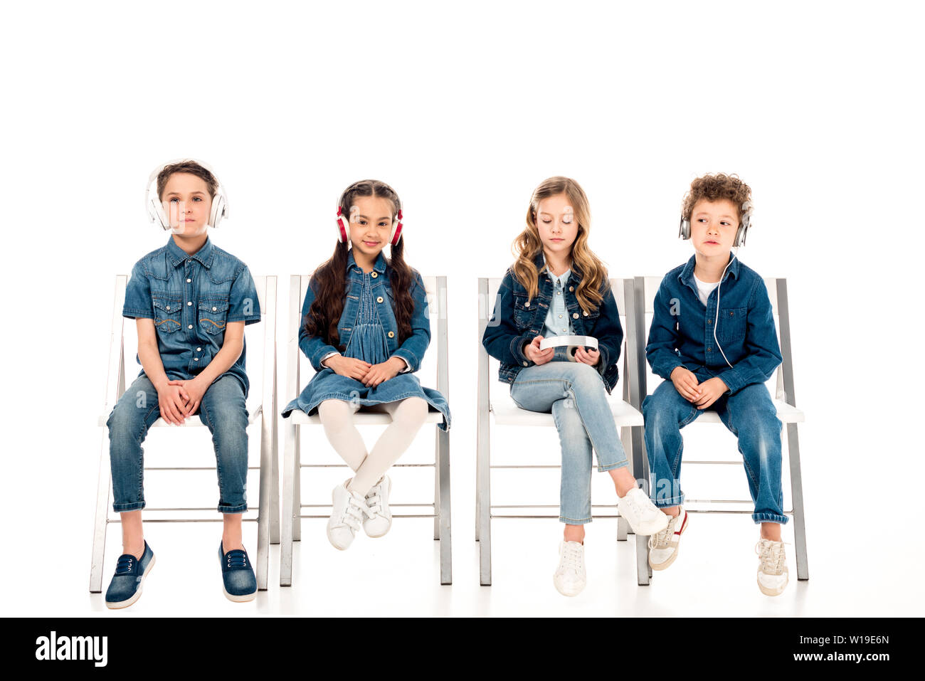 Vier Kinder in Jeans Kleidung sitzen auf Stühlen und das Hören von Musik über Kopfhörer auf Weiß Stockfoto