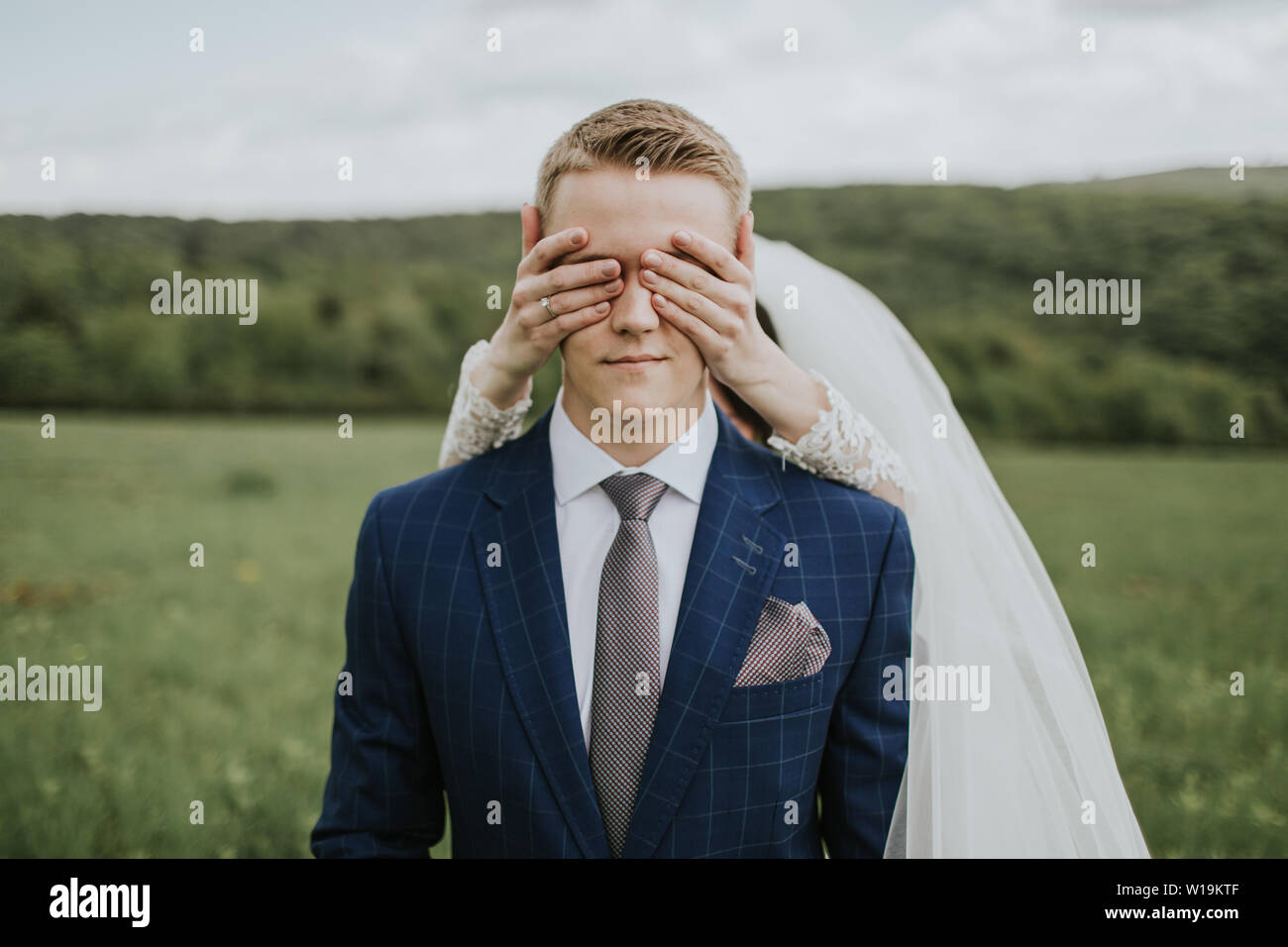 Braut, die Augen des Bräutigams. Portrait der junge Bräutigam wartet auf Wiese und Braut, die hinter ihm stehen, die seine Augen. Stockfoto