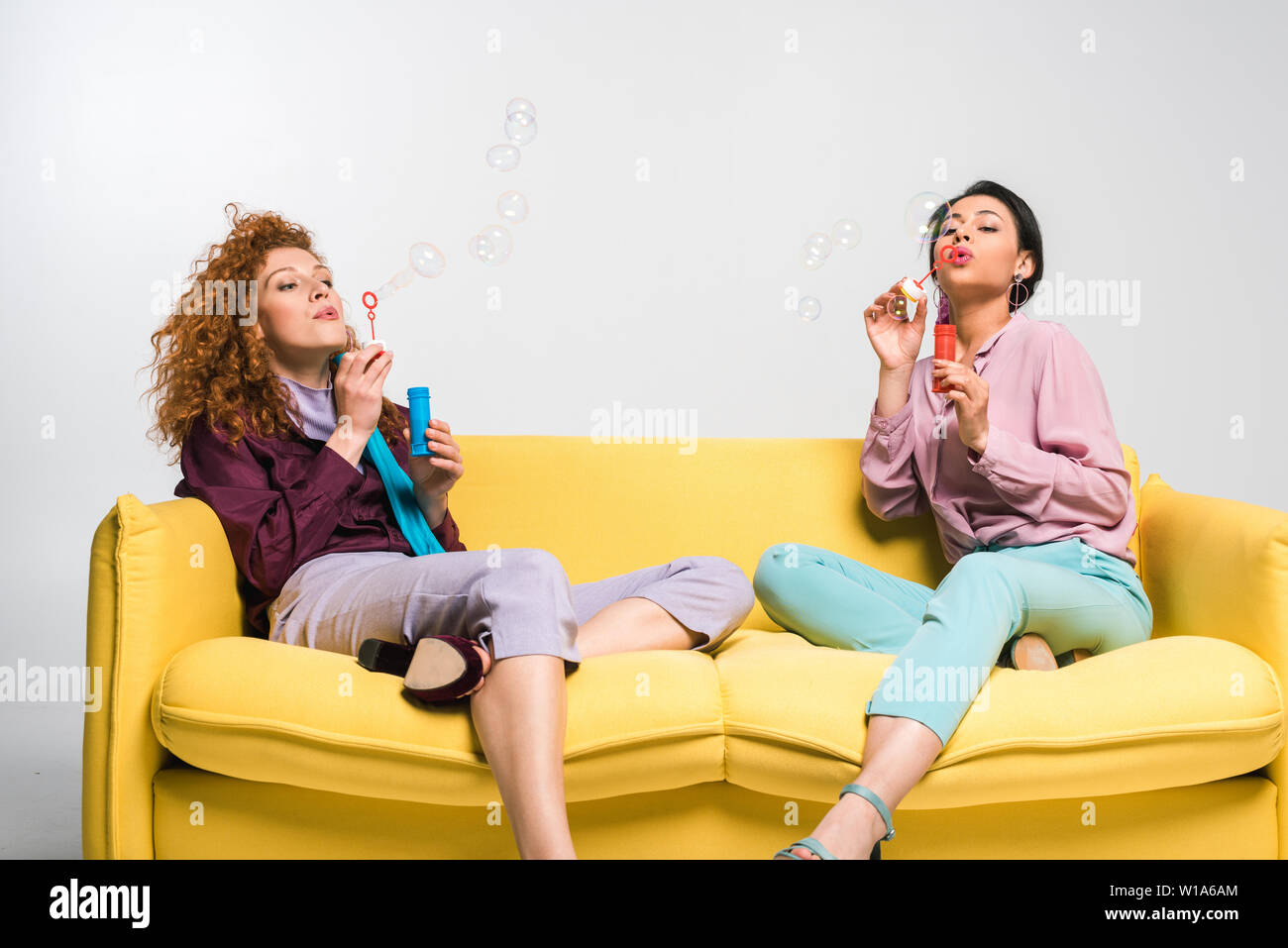Curly rothaarige Mädchen und afrikanische amerikanische Frau bläst Seifenblasen beim Sitzen auf einem Sofa gelb auf weiß Stockfoto