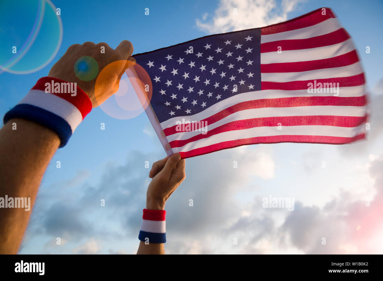 Hände mit USA roten, weißen und blauen Armbänder Holding eine amerikanische Flagge schwenkten in hellen Himmel mit sonnigen lens flare Stockfoto
