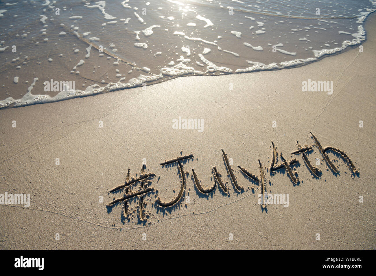 Moderne Nachricht für den 4. Juli amerikanischen Feiertag mit einem Social Media - freundliche hashtag in glatten Sand geschrieben mit ankommenden Welle auf den Strand Stockfoto