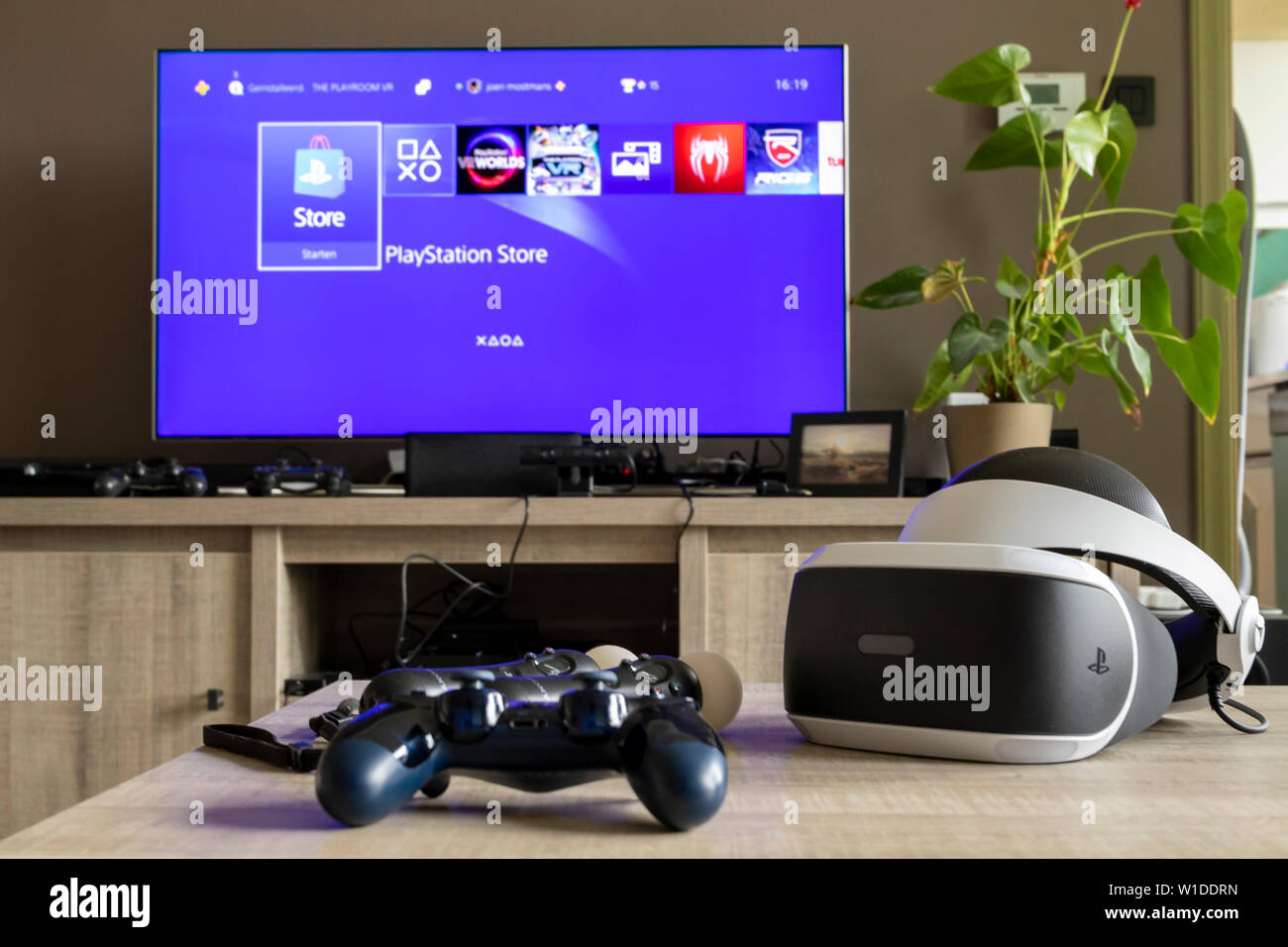 Eine playstation VR-Headset an einem hölzernen Tisch zusammen mit einer  PS4-Steuerung und einige Playstation Move Controller. Im Hintergrund gibt  es einen Fernseher Stockfotografie - Alamy