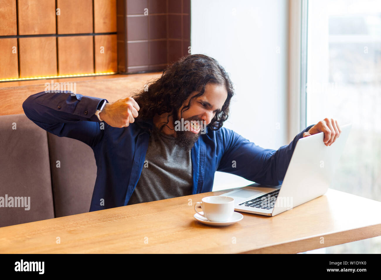 Portrait von Angry aggressiver junger erwachsener Mann Freiberufler in lässigen Stil im Cafe sitzen und versuchen, Laptop Bildschirm Absturz, erhobene Faust mit Zusammenpressen Stockfoto