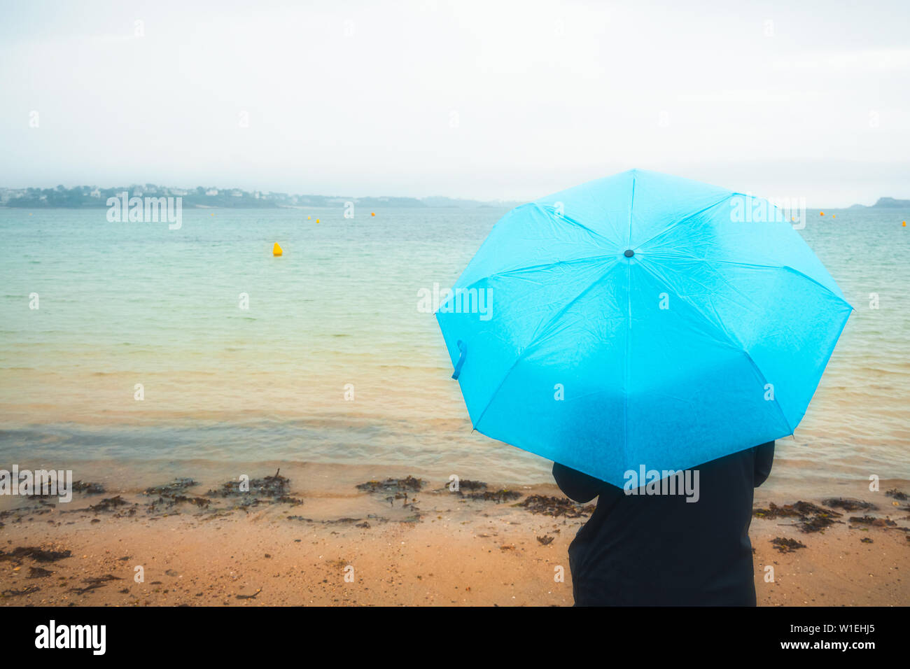 Zurück oder hinten Blick auf eine Frau, die durch das Meer in Regenjacke und mit einem blauen Schirm an einem regnerischen und Moody Tag. Wetter Konzept mit kopieren. Oran Stockfoto