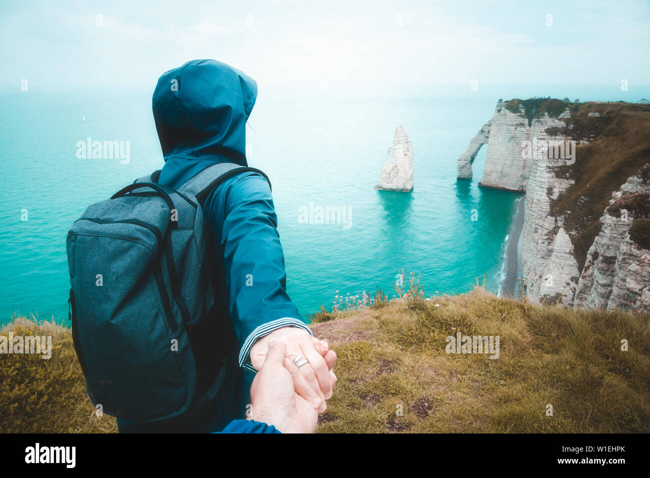 Folgen Sie mir. Junge Frau in Blau Regenjacke die Hand von ihrem Freund zu Fuß bis an den Rand der Klippe, einer malerischen Seenlandschaft zu beobachten, Etretat, Frankreich. Stockfoto