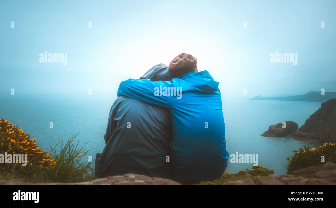 Ansicht von hinten oder hinten Blick auf zwei junge Erwachsene Liebhaber sitzen auf dem Rand einer Klippe auf das Meer an einem regnerischen und Moody Tag. Partnerschaft, Draußen, tra Stockfoto