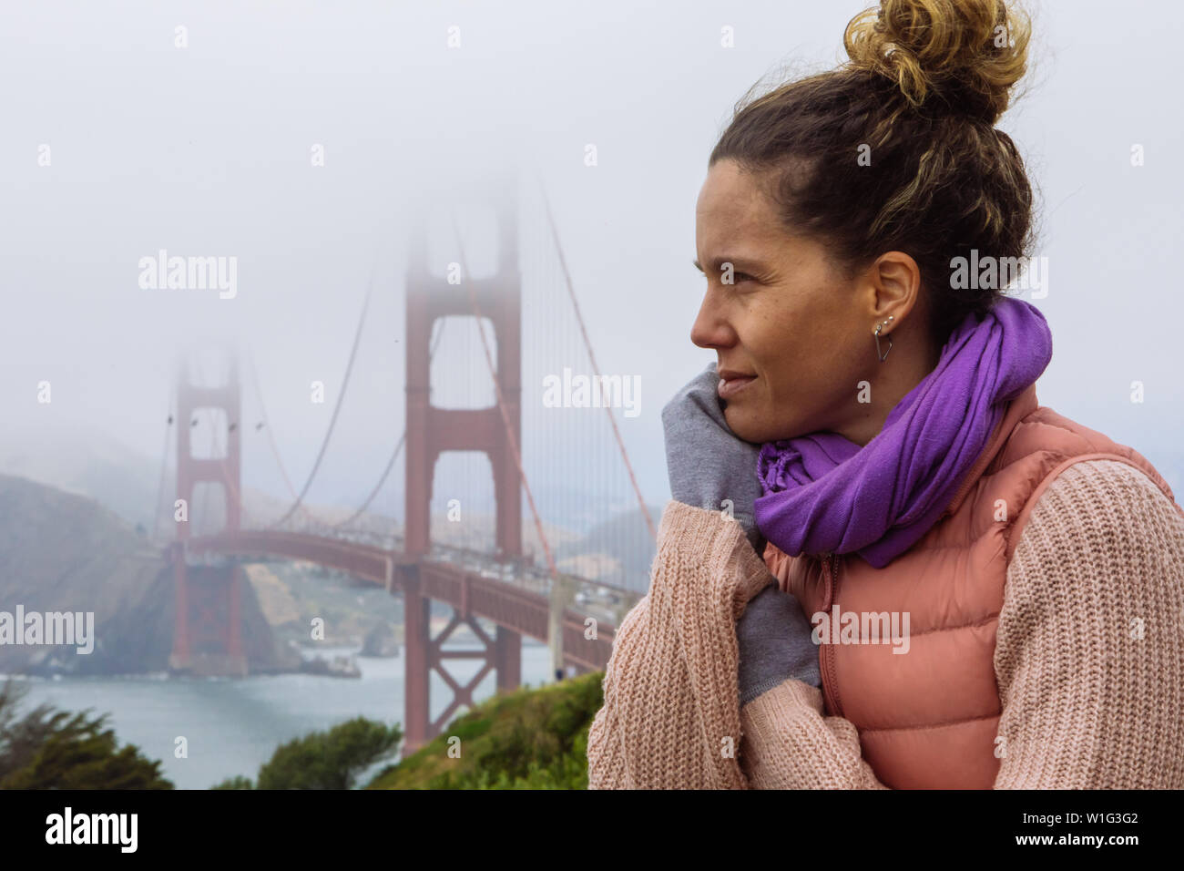 Lockiges Haar kaukasische Frau in den Dreißigern mit einem Brötchen, das mit der Golden Gate Bridge in San Francisco, Kalifornien, USA, posiert Stockfoto
