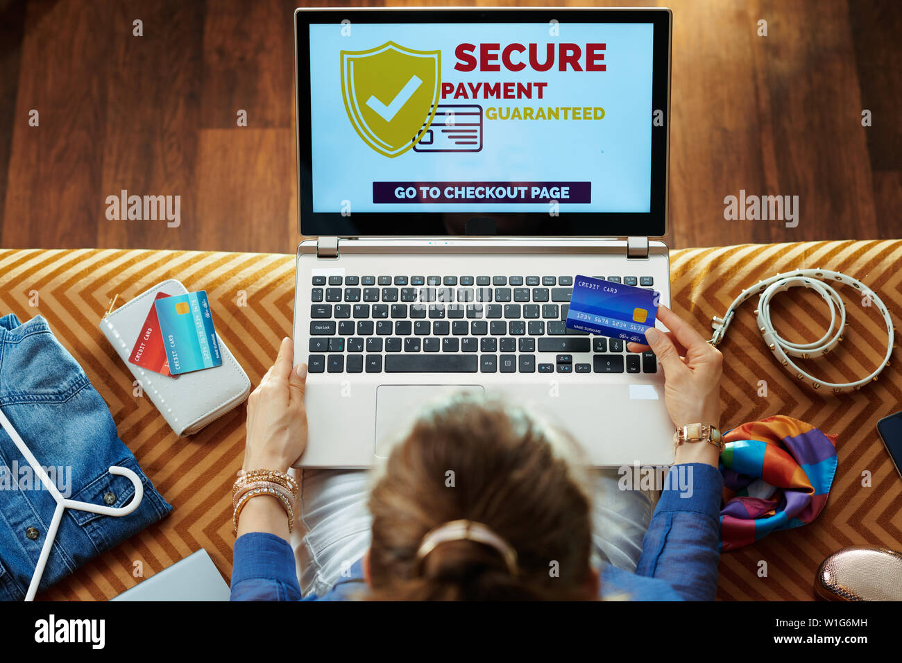 Obere Ansicht von stilvollen Frau in Blau Bluse mit Kreditkarte kaufen sicher im Internet auf einem Laptop sitzend auf Schlafcouch im Wohnzimmer. Stockfoto