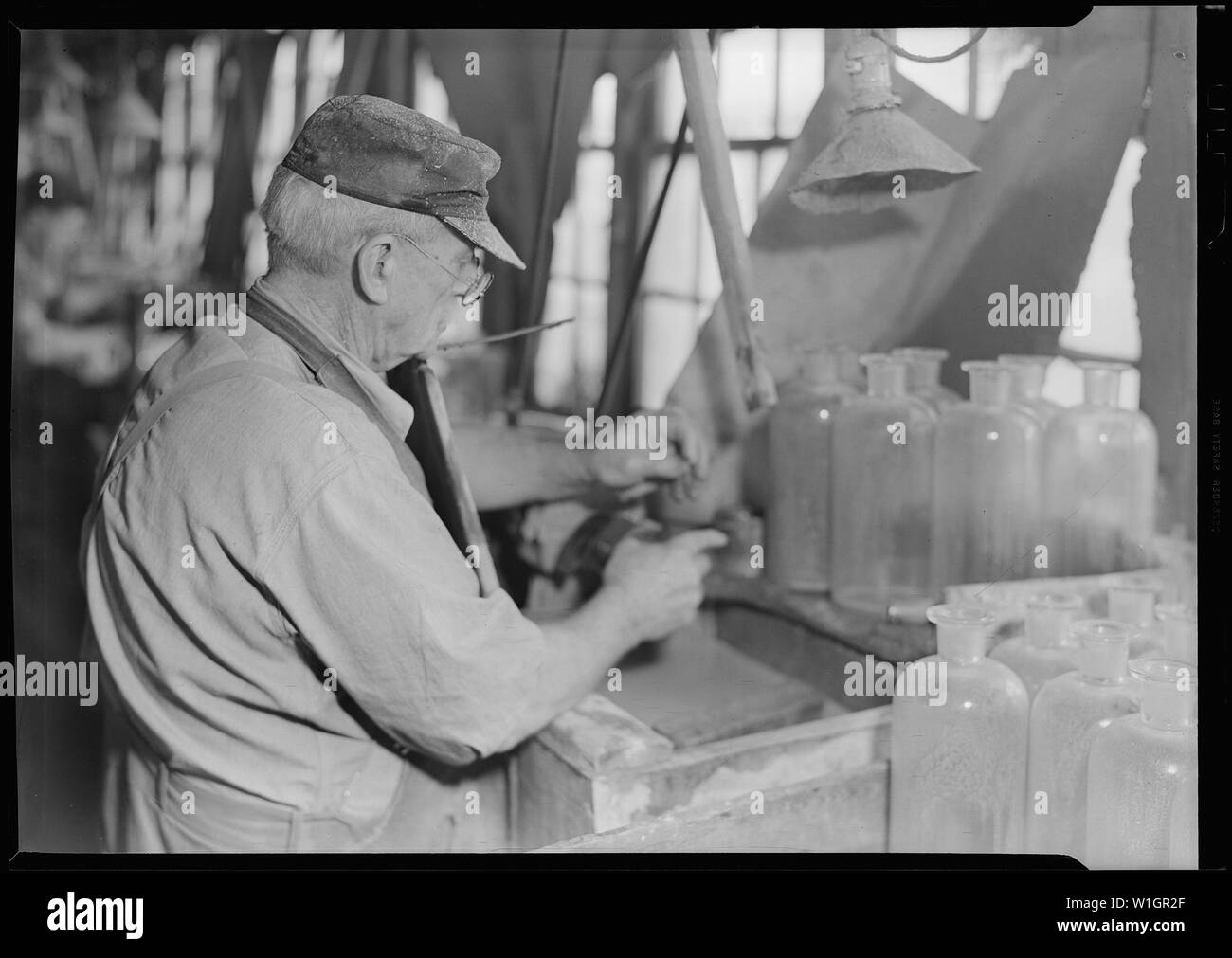 Millville, New Jersey - Glasflaschen. Stopper - Schleifer an T.C. Wheaton Co; Umfang und Inhalt: Millville, New Jersey - Glasflaschen. Stopper - Schleifer an T.C. Wheaton Co.Dieser Mann 83 Jahre alt für diese Fabrik an diesem Job gearbeitet hat, seit er 15 war. Er ist der Glättung und Gestaltung Stopfen die Flaschen an der rechten unteren Ecke des Bildes zu passen. Mit einer Lösung von Bimsstein und Wasser er benetzt die Emery Rad genau die Form der Hals der Flasche. Er wirbelt die Emery Rad durch Drücken auf den Fuß tritt. Durch Drücken auf den Stöpsel der Flasche gegen diese Emery Rad ist er in der Lage zu Stockfoto