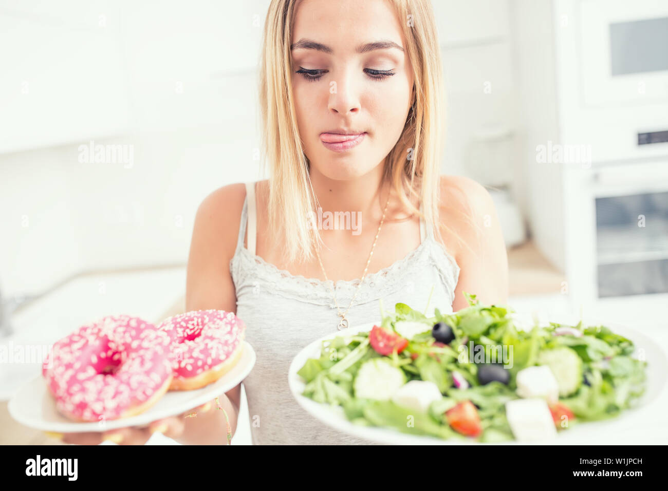 Junge blonde Mädchen beim Frühstück oder Abendessen in der Küche zu Hause wählt zwischen Donut und Salat. Stockfoto