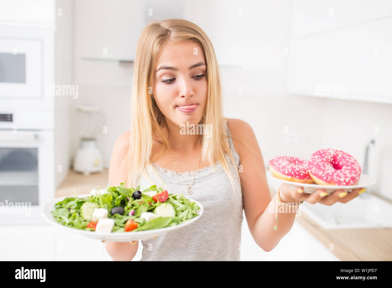 Junge blonde Mädchen beim Frühstück oder Abendessen in der Küche zu Hause wählt zwischen Donut und Salat. Stockfoto