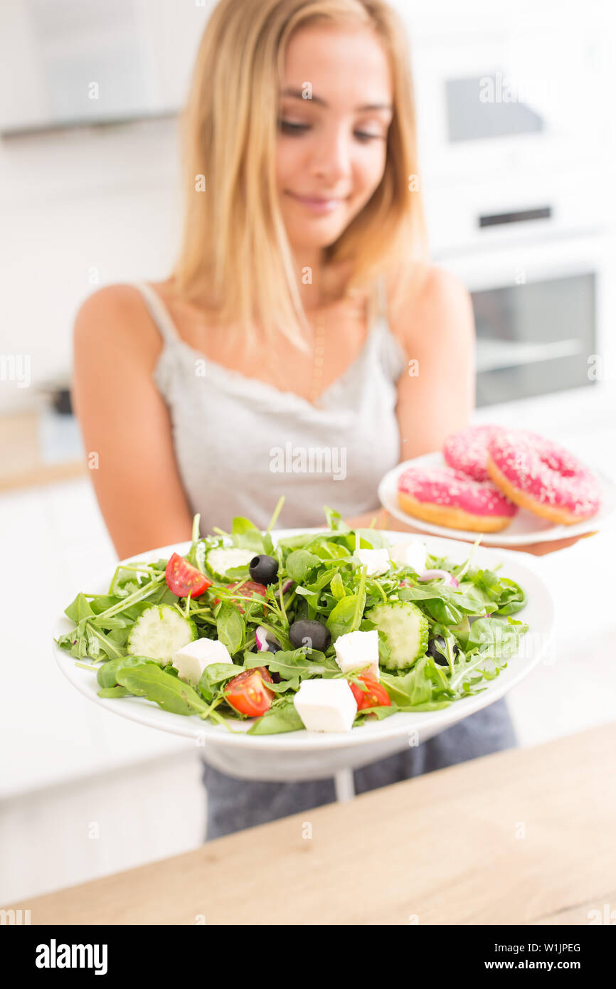 Junge blonde Mädchen beim Frühstück oder Abendessen in der Küche zu Hause wählt zwischen Donut und Salat. Stockfoto