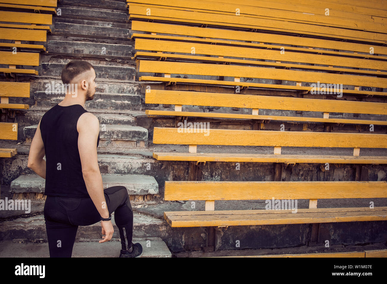 Ein schlanker junger Mann in Schwarz Sportswear steht vor der Treppe Stadion steht. Einsatzbereit, Schritt zum Sieg, Herausforderung. Stockfoto