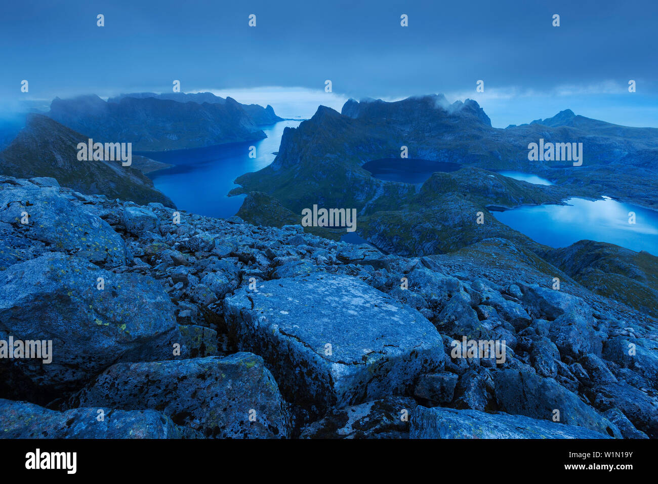 Blick von der Kante des Hermannsdalstinden im Blau der Dämmerung auf die umliegenden Berge mit Seen und den Kjerkfjorden in der backgro Stockfoto