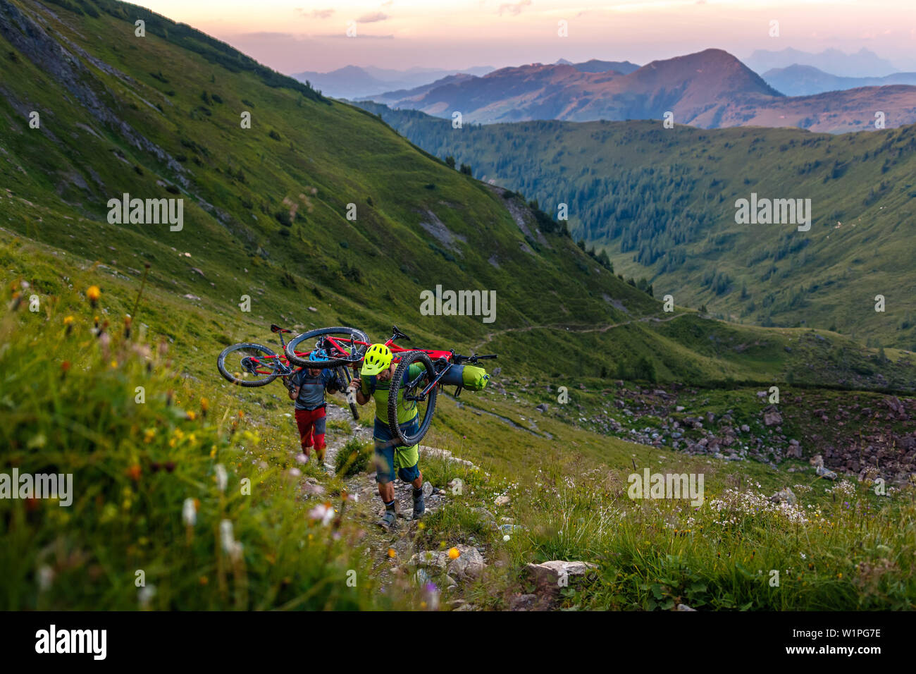 Zwei Mountainbiker tragen ihre Bikes auf der Rückseite, nach Sonnenuntergang, grüne Gras bewachsenen Berghang, Kirchberg, Tirol, Österreich Stockfoto