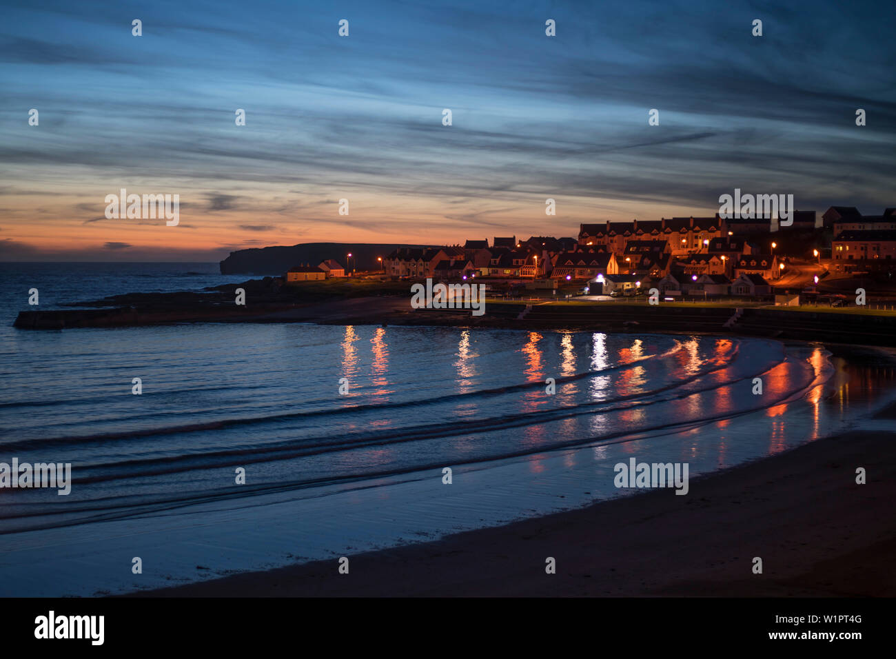 Die Lichter der Stadt spiegeln sich im Wasser des Atlantischen Ozeans in Kilkee Bay unter einer rosa Himmel bei Sonnenuntergang, Kilkee, County Clare, Irland, Europa Stockfoto