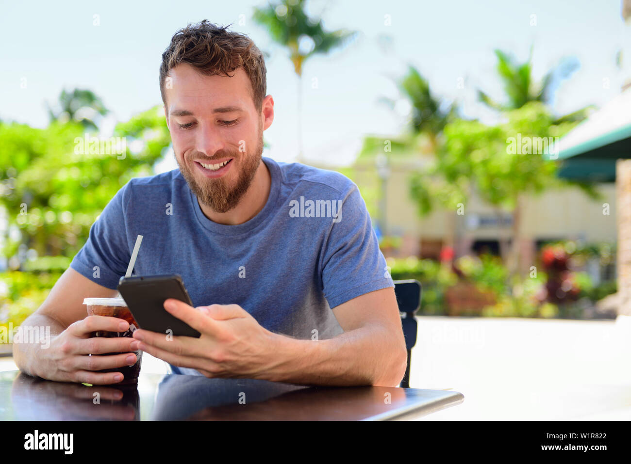 Smartphone Mann sms Trinken kalten Kaffee trinken im Café im Freien auf der Terrasse im Sommer. Happy and Urban junger Erwachsener holding Telefon lesen oder mit "App mit Freunden über soziale Medien zu verbinden. Stockfoto