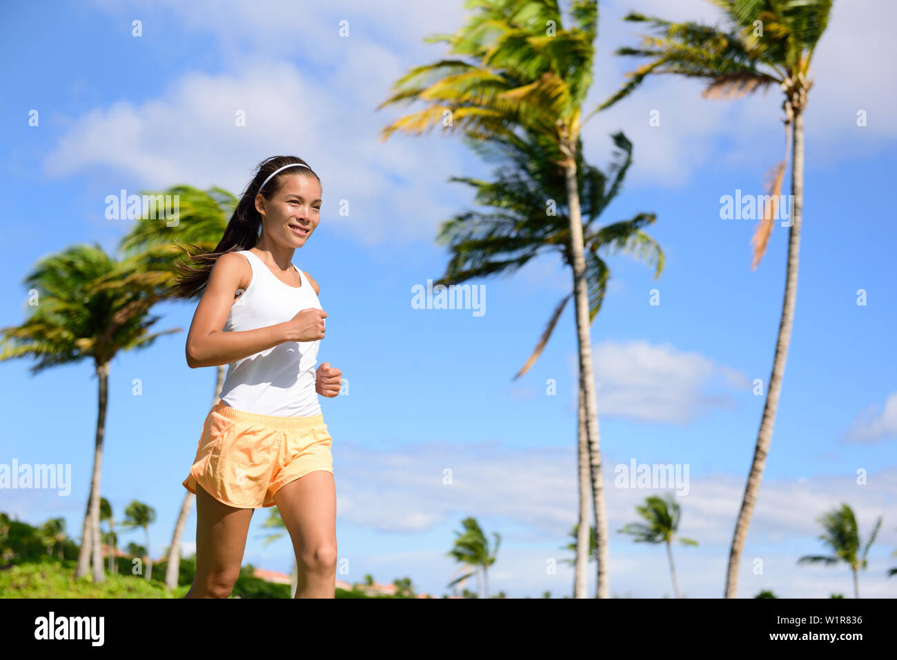 Asiatische runner girl Jogging in tropischen Natur Park im Freien mit Palmen Hintergrund im Sommer. Junge Erwachsene sport Frau Laufen fit tragen orange Fitness Shorts und Tank Top. Stockfoto