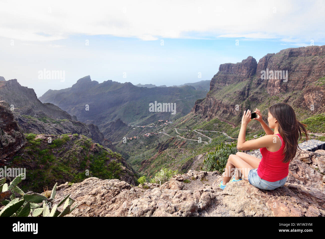 Frau fotografieren Berge beim Sitzen auf einer Klippe. Junge weibliche wird mittels Smart Phone klicken Sie auf das Bild einer schönen Natur. Rückansicht Tourist besuchen Teneriffa Masca Tal, Kanarische Inseln, Spanien. Stockfoto