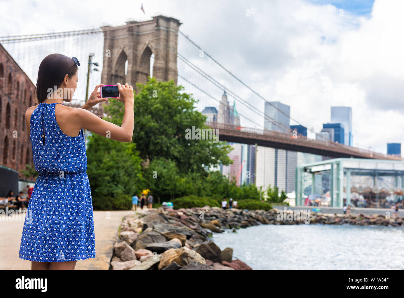 Tourist, travel Bild mit Telefon der Brooklyn Bridge und die Skyline von New York City während der Sommerferien. Nicht erkennbare weiblichen jungen Erwachsenen genießen USA Ferien in blauem Kleid. Stockfoto
