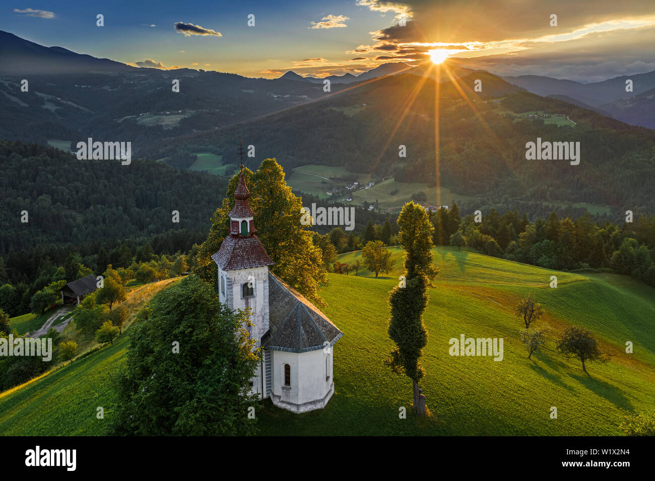 Skofja Loka, Slowenien - Luftbild der schönen Hügel Sveti Tomaž (Hl. Thomas) Kirche mit einem herrlichen Sonnenuntergang und die Julischen Alpen im backgroun Stockfoto