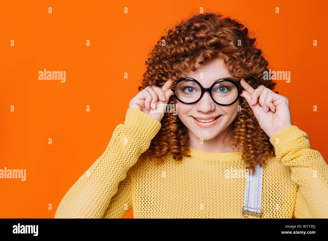 Curly - behaarte Frau ihre Brille ajusting auf orange Hintergrund. tennage Mädchen mit stilvollen hipster Gläser Stockfoto