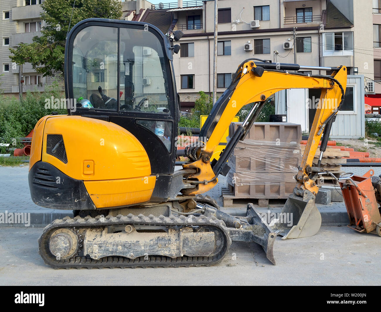Gelbe Mini Bagger an den Anschlüssen für kleine Bauarbeiten an schwer zugänglichen Stellen oder auf schmalen Straßen der Stadt, Seitenansicht, Sommertag. Stockfoto