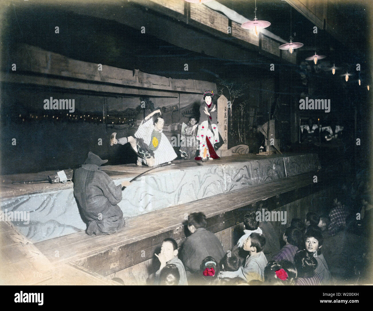 [1900s Japan - Japanische Theater Performance] - Schauspieler auf einer Bühne vor einem Publikum von Kindern in einem Theater. Ein "kuroko's' (Stufe Assistant in Schwarz gekleidet) können in der vorderen gesehen werden. Seinen Kopf bis Fuß schwarze Kleidung impliziert seine Unsichtbarkeit auf der Bühne. Auf der rechten Seite die Umrisse der Musiker bei Kerzenlicht gesehen werden kann. 20. jahrhundert Vintage albumen Foto. Stockfoto