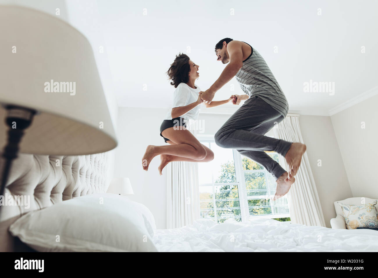 Mann und Frau halten sich an den Händen und springen auf dem Bett. Paar Spaß haben im Schlafzimmer. Stockfoto