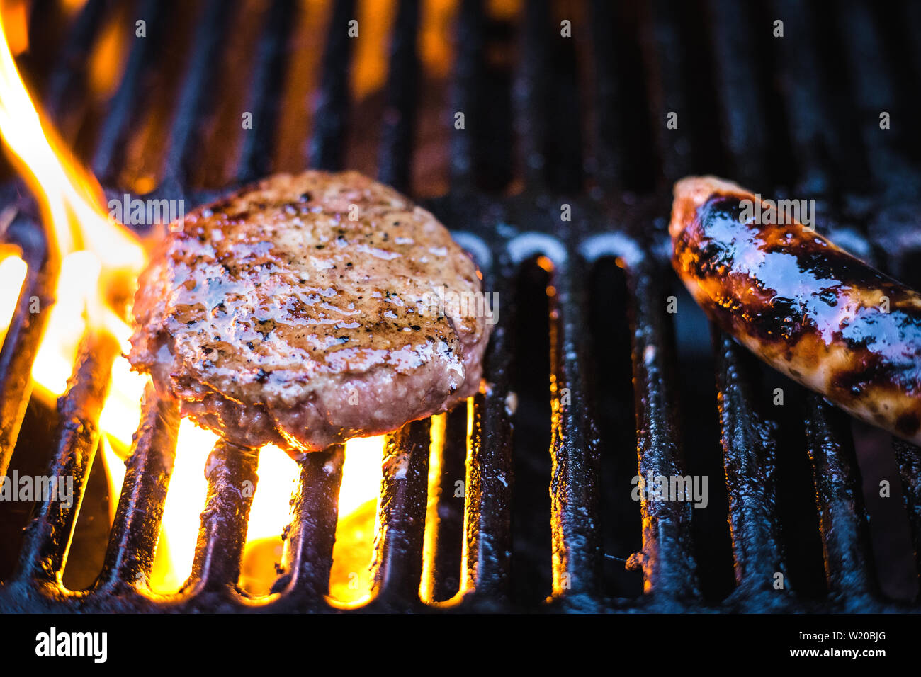 Sommer kochen auf einem Grill mit Beef Burger und Würstchen. Flammen sind Aufflammen um eine teilweise gekochtes Rindfleisch Burger. Stockfoto