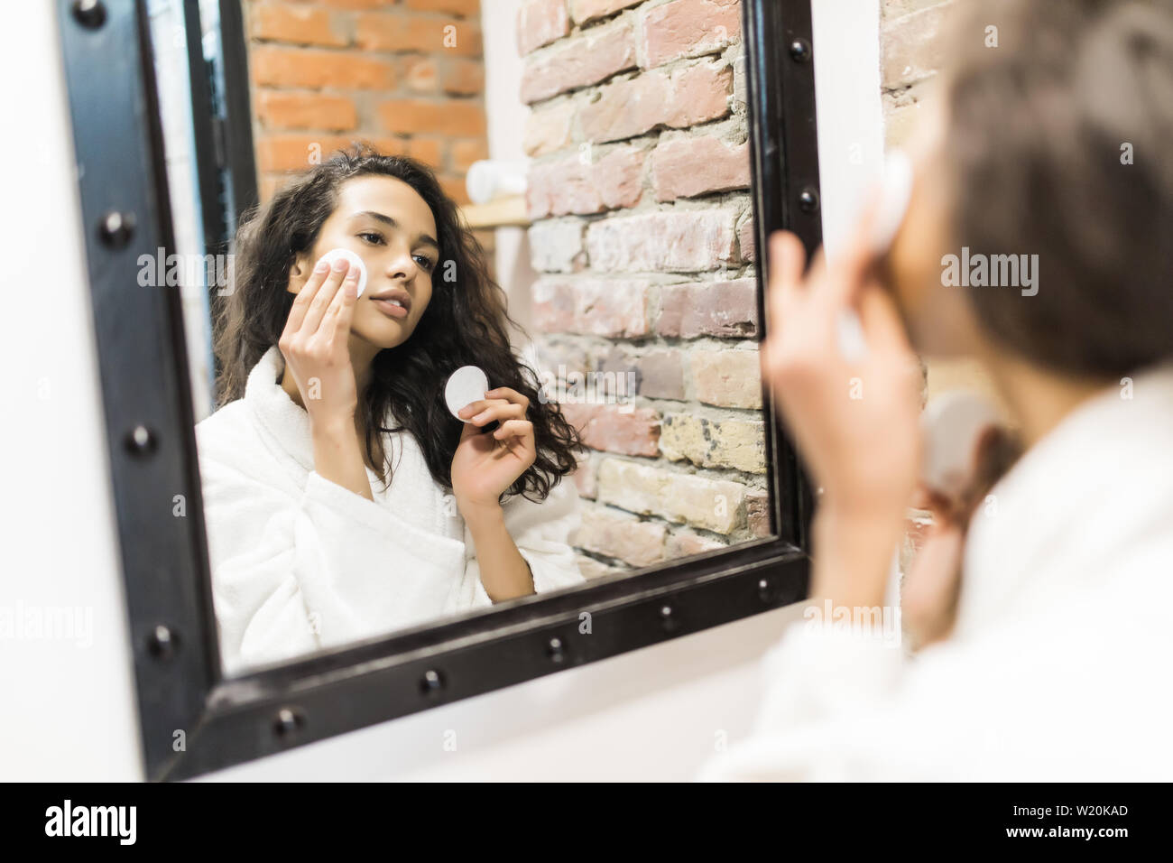 Schöne junge Frau ist Reinigung Ihr Gesicht mit einem Baumwolle Disc und lächelnd in den Spiegel im Badezimmer suchen Stockfoto