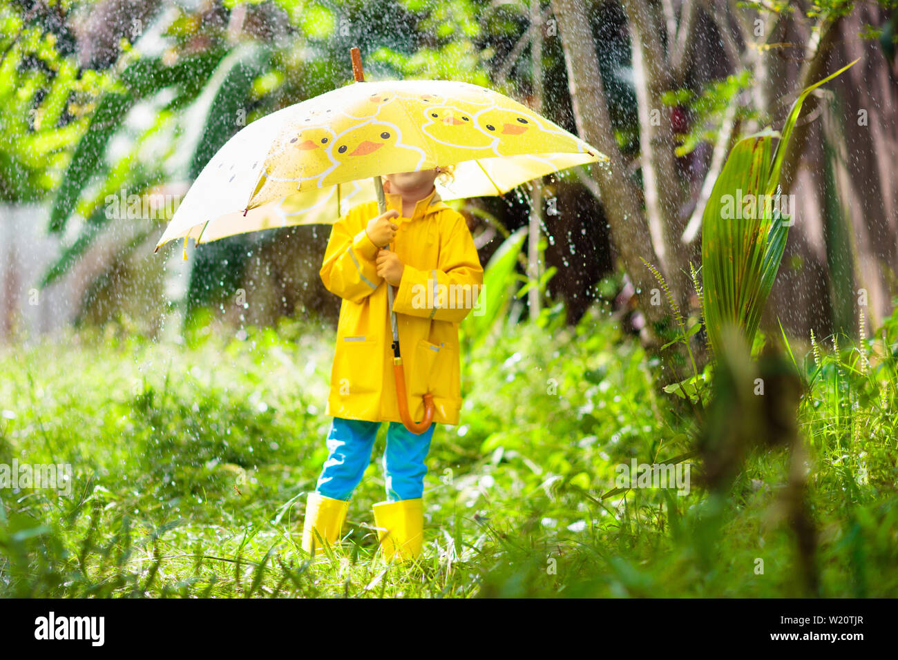 Kinder spielen im Regen an sonnigen Herbsttag. Kind unter schweren Dusche mit gelber Ente Regenschirm. Kleine Junge mit Entlein wasserdichte Schuhe. Gummi wel Stockfoto