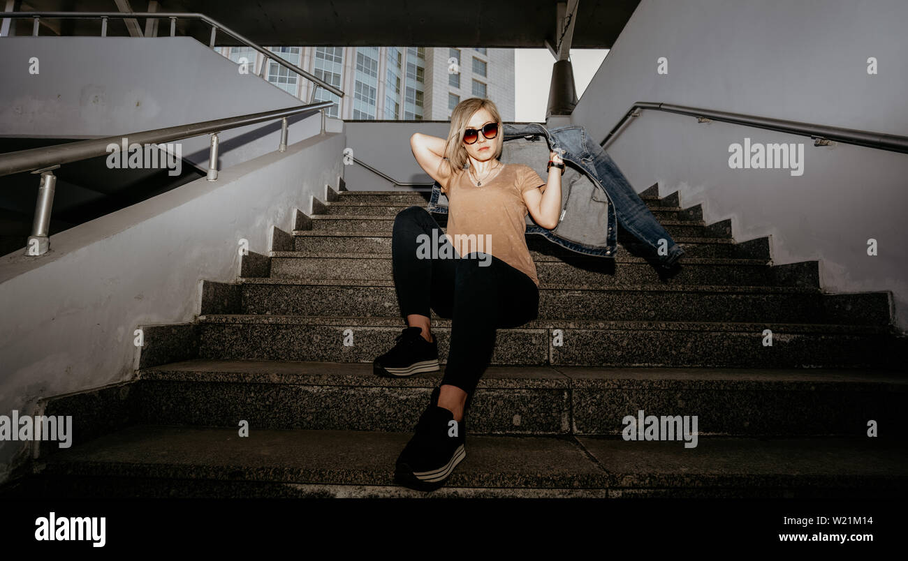 Blondine in Sonnenbrille trägt eine Jeansjacke sitzt auf den Stufen. Moderne Mädchen. Stockfoto