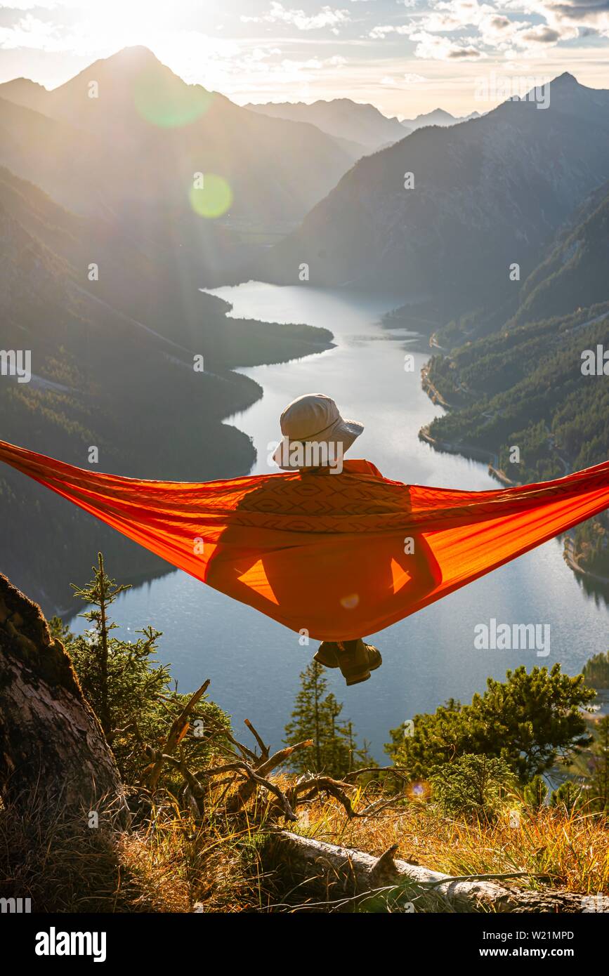 Mann mit einem Sonnenhut in einem orange Hängematte sitzen, Panoramablick auf Berge mit See, Plansee, Tirol, Österreich Stockfoto