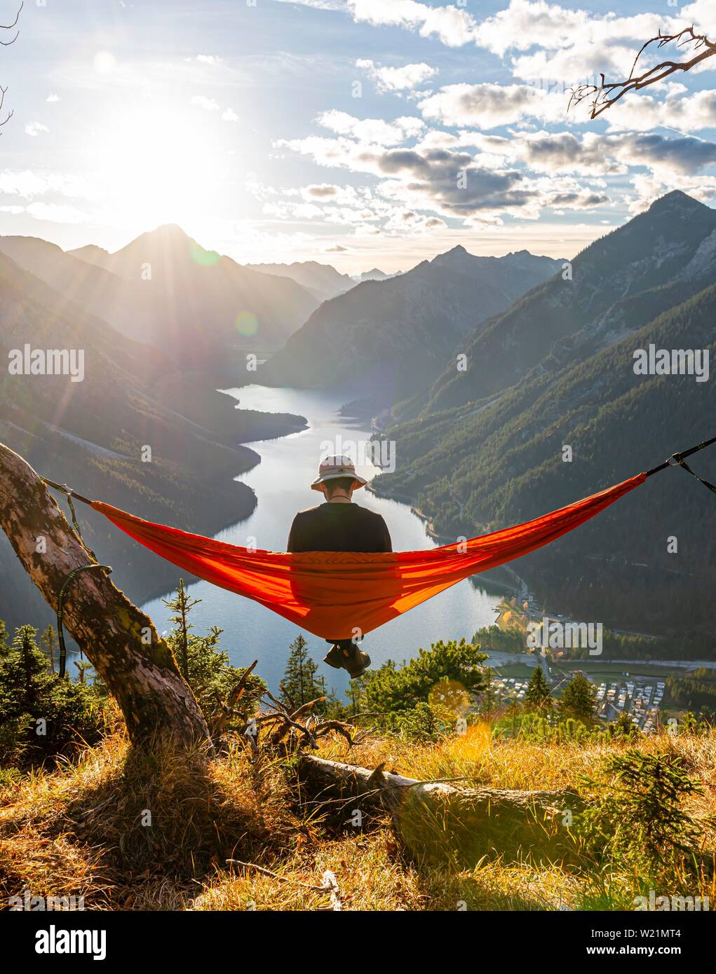 Mann mit einem Sonnenhut in einem orange Hängematte sitzen, Panoramablick auf Berge mit See, Plansee, Tirol, Österreich Stockfoto