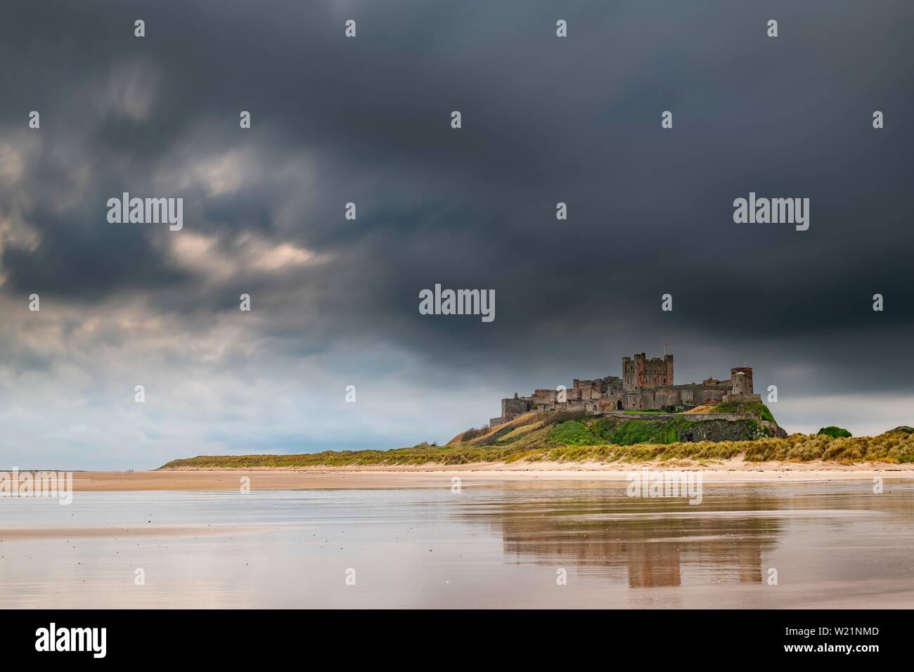 Flache sandige Küste mit Bamburgh Castle und Spiegelbild im Wasser mit dunklen Wolken, Bamburgh, Neue castele upon Tyne, Northumberland, Großbritannien Stockfoto