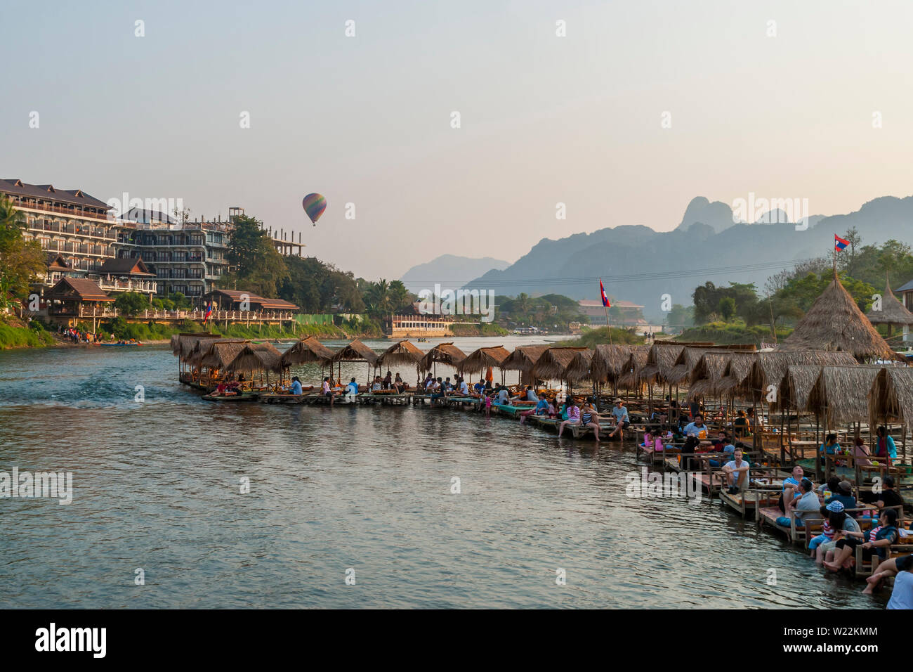 Vang Vieng, Laos - Feb 2016: Personen, die bei einer schwimmenden Bar am Fluss in Vang Vieng und Heißluftballon über der Stadt fliegen Stockfoto