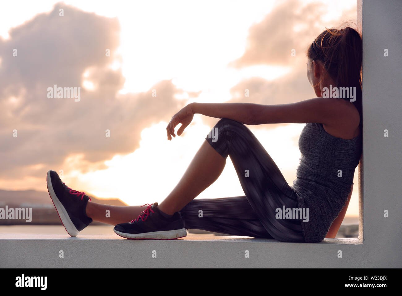 Fitness Athlet sport Frau Entspannung nach dem Training sitzen auf Fenster mit Aussicht auf den Sonnenuntergang. Silhouette der Läufer in Activewear zu Hause auf der Terrasse im Freien Himmel Hintergrund. Stockfoto