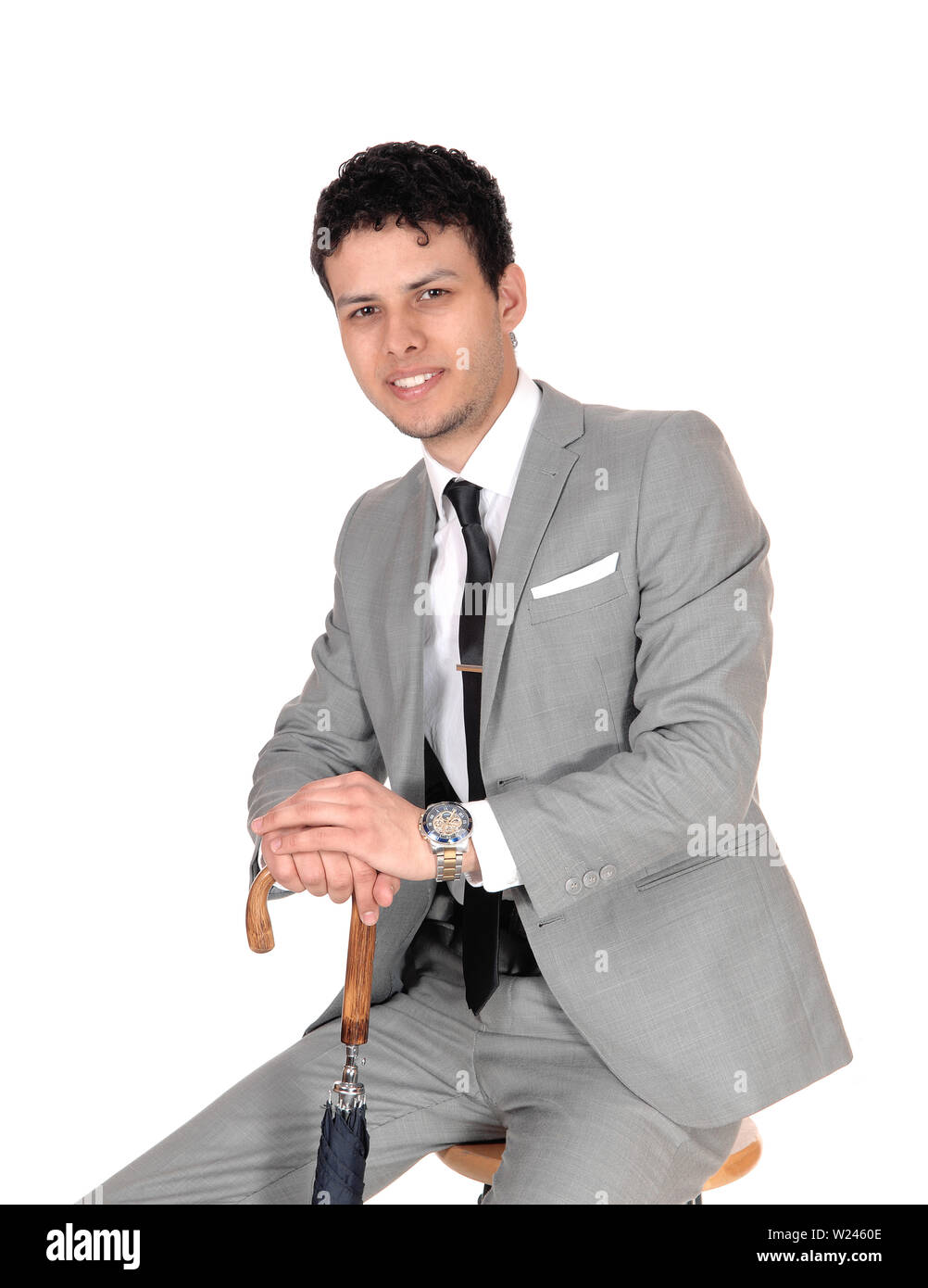 Ein junger Mann in einem grauen Anzug sitzt in einem Studio für weissen Hintergrund mit seinen Händen auf einen Schirm und lächelnd Stockfoto