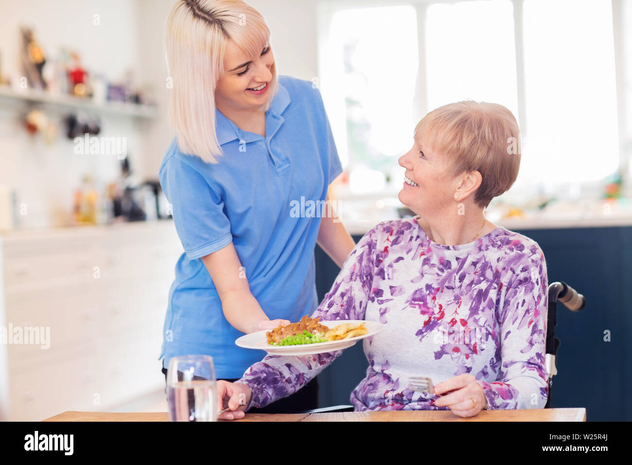 Weibliche Care Assistant servieren Mahlzeit zu älteren Frau sitzt im Rollstuhl am Tisch Stockfoto