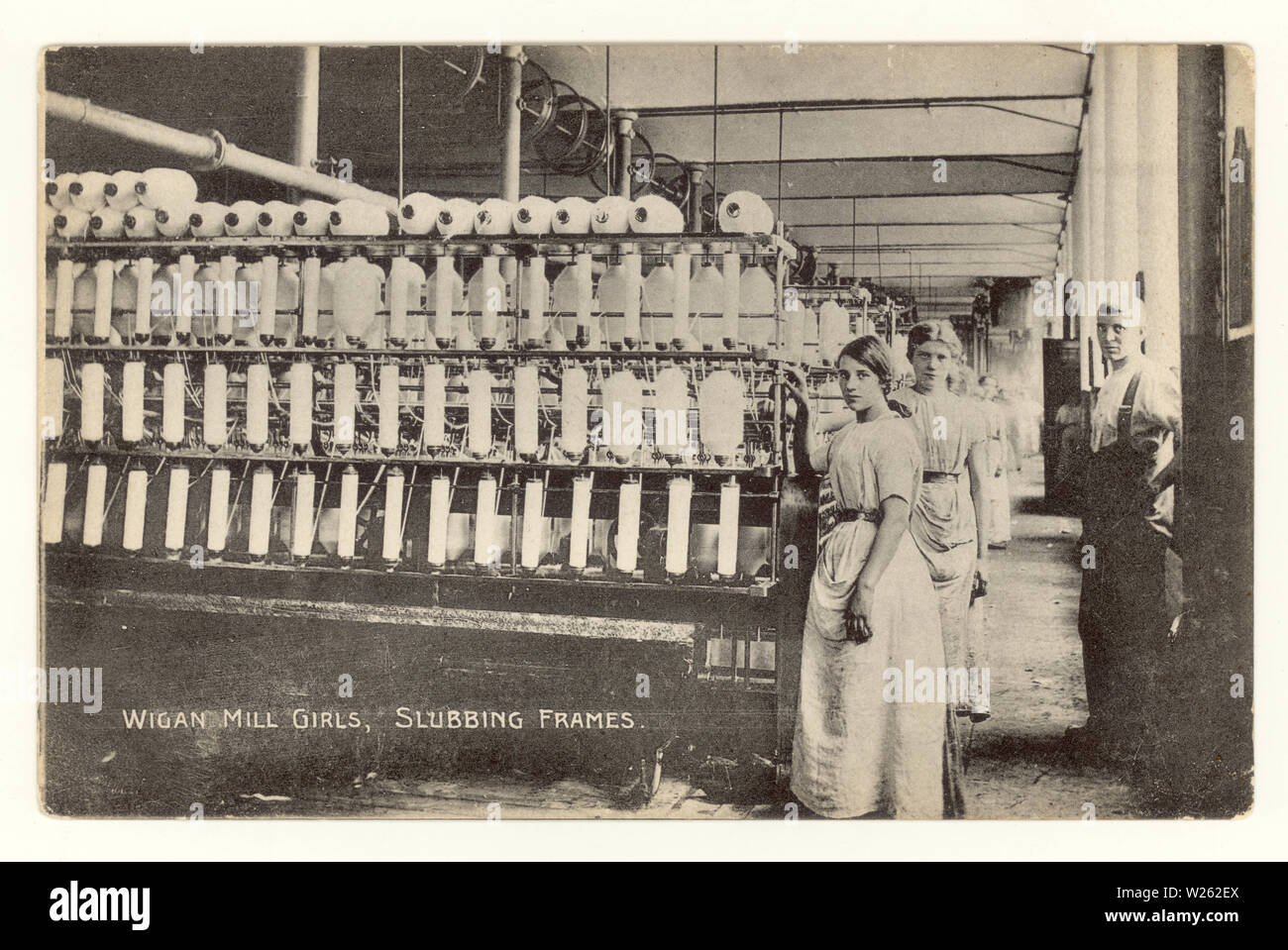 Frühe Postkarte unten gedruckt wird 'Wigan Mühle Mädchen, slubbing frames', mit Aufseher/Meister stand neben slubbing Frames - eine Maschine verwendet, um die Fasern in der Baumwolle zu begradigen vor spinning 'Lancashire Lasses', um 1910, Wigan, Greater Manchester, Großbritannien Stockfoto