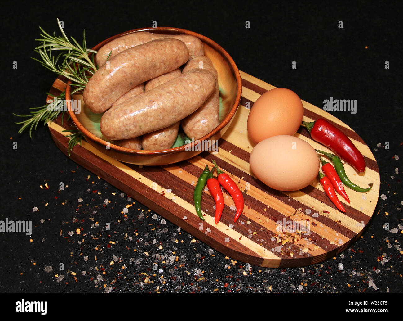 Frische Würstchen in einer Schüssel auf einem Holzbrett mit Eier und rote Chilischoten Stockfoto