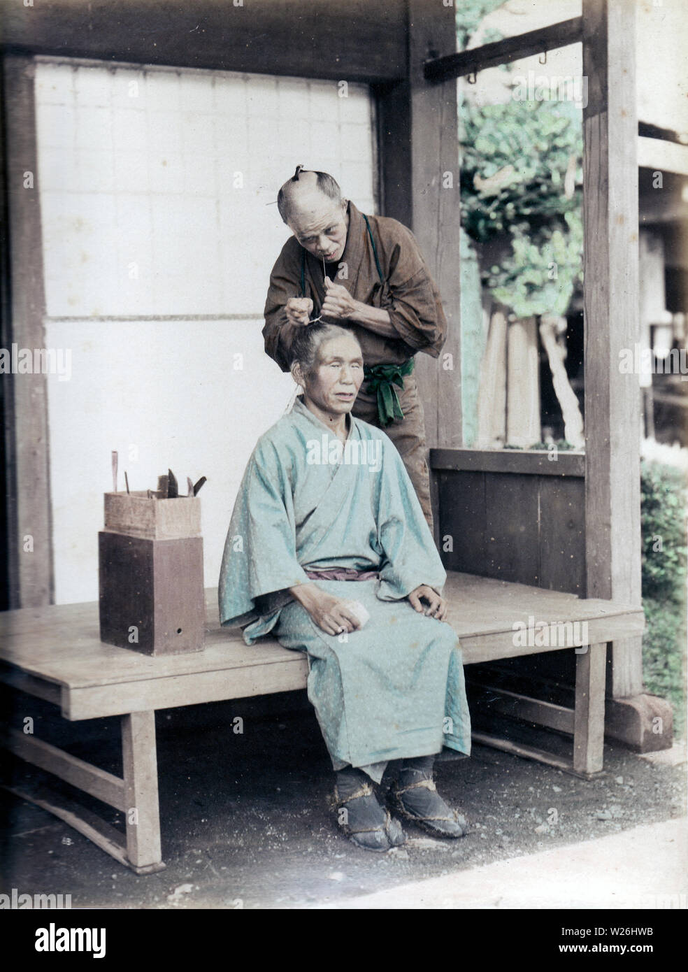 [1890s Japan - Japanische Friseur bei der Arbeit] - ein Friseur die Haare eines männlichen Kunden. Bis westlichen Frisuren während der Meiji Periode (1868-1912) gefördert wurden, japanische Männer trug ein chonmage Haarschopf. Der Werkzeugkasten, deutet darauf hin, dass Dies ist ein Reisen Friseur. 19 Vintage albumen Foto. Stockfoto