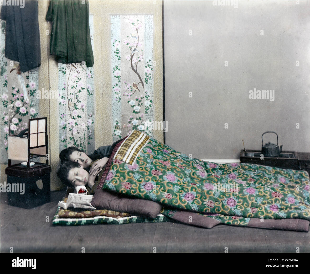 [1890s Japan - Japanische Frauen Schlafen] - Ein studio Foto von zwei Frauen in wattierte Futon Betten schlafen mit dem Kopf auf hölzernen Kopf unterstützt. Es muss im Winter sein, da sie zwei Schichten sowohl für die Matratze und Abdeckung verwenden. Es ist ein Ando Lampe in der Nähe von ihren Köpfen. Ein Paravent mit Blumen und Vögel illustriert steht dahinter. Die in der Nähe ihrer Füße ist ein hakohibachi (brazier in einer Holzkiste verpackt) mit einem tetsubin (Bügeleisen, Wasserkocher). 19 Vintage albumen Foto. Stockfoto