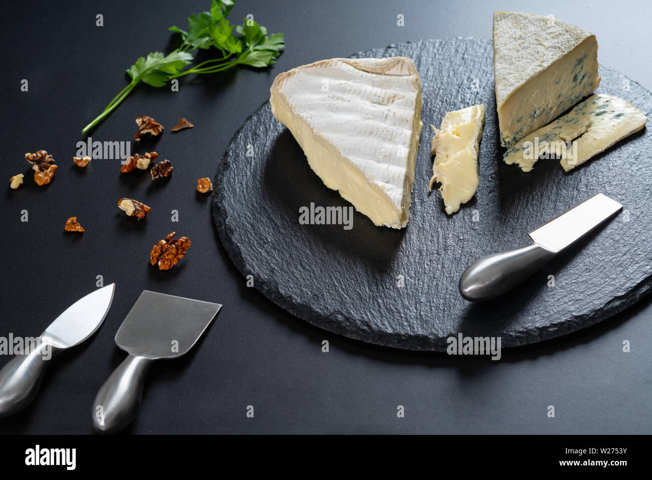 Keile von semi-soft Käse auf einem schwarzen Holz- Käseplatte mit Stahl Cutter und zerstreute gehackte Walnüsse in einem hohen Winkel food Still Life Stockfoto