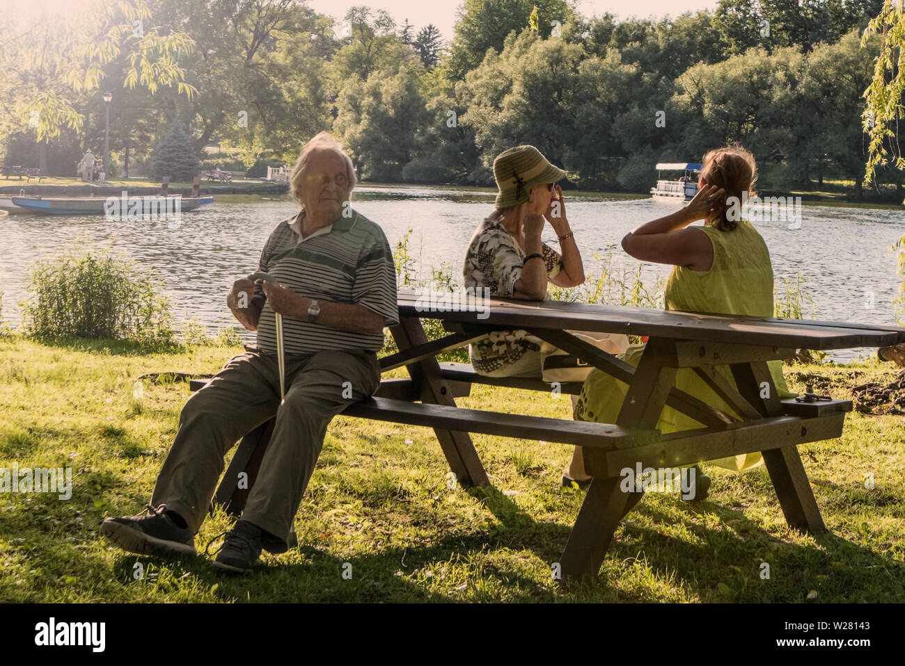 Zwei älteren Damen und ein alter Mann sitzt auf einer Picknickbank, die Frauen Interaktion, der ältere Mann hat einen Stock, und er ist das Ignorieren der Damen. Stockfoto