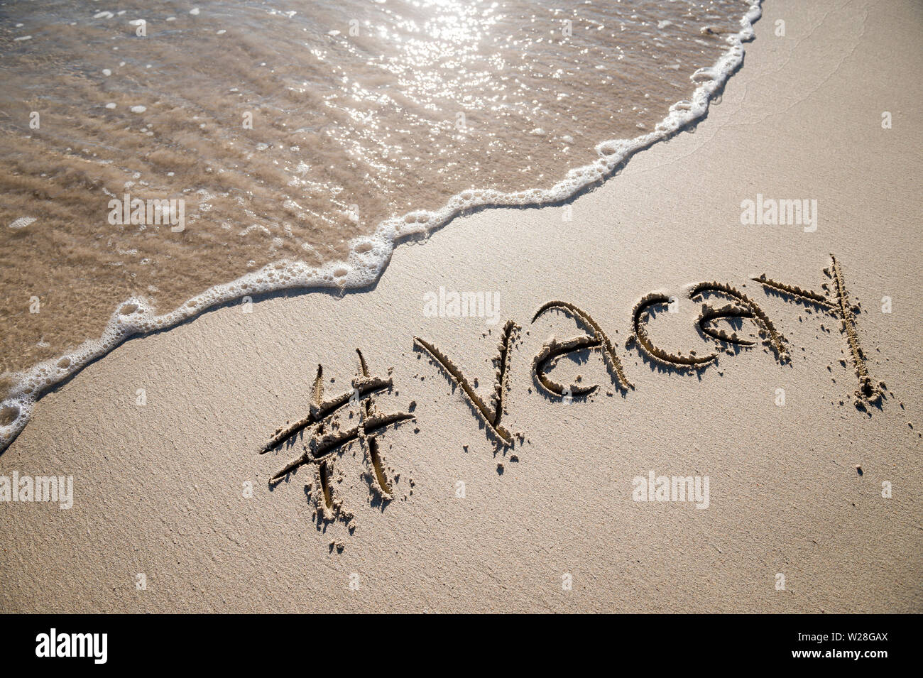 Modernes reisen Nachricht für den Strand mit einem Social Media - freundliche Hashtag mit dem Wort "vacay" in sanften Sand mit ankommenden Welle geschrieben Stockfoto