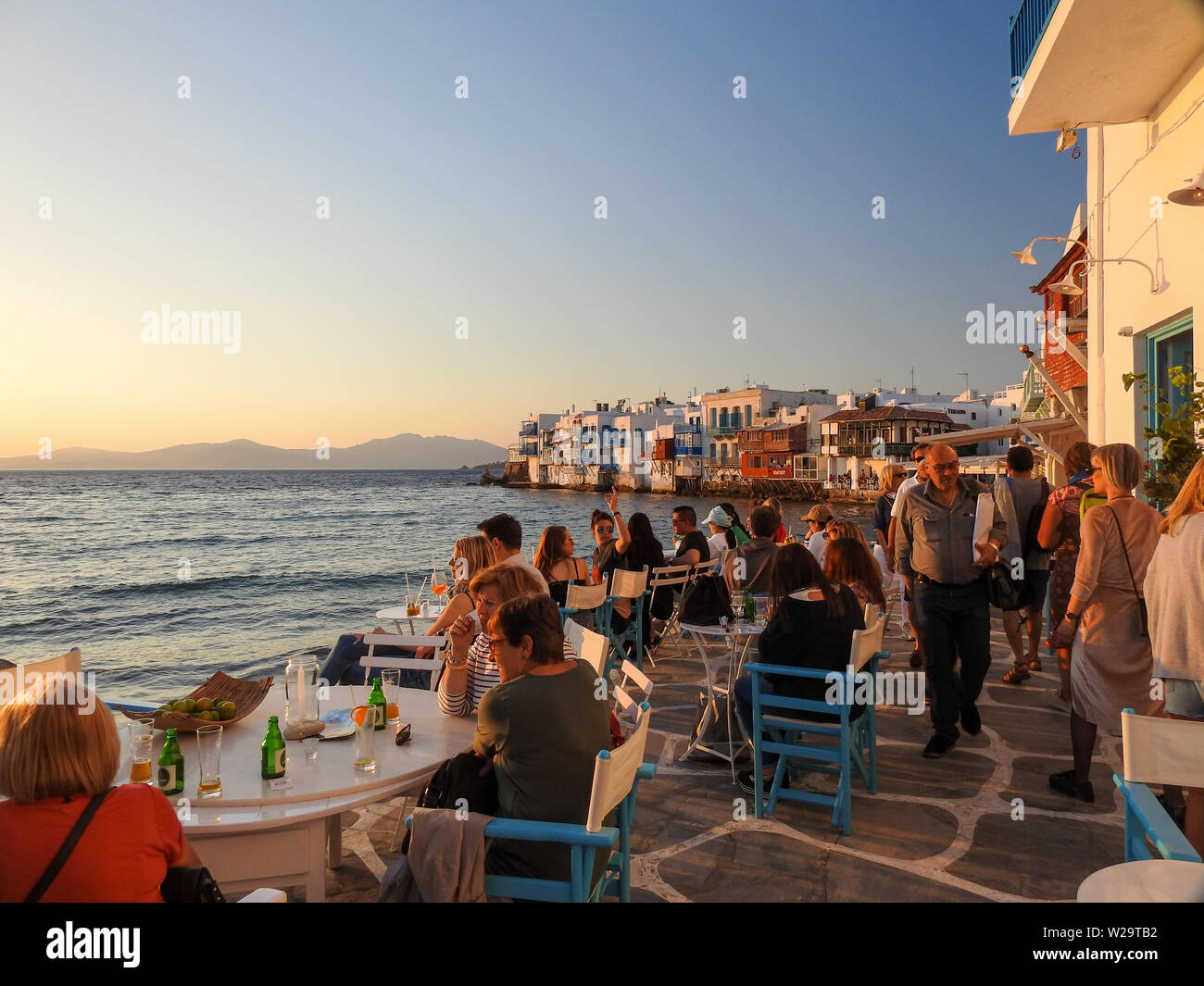 Mykonos, griechische Inseln, Griechenland die Menschen essen im Freien auf goldenen sonnigen Nachmittag mit weißen und blauen Gebäuden und Wasser, Stockfoto