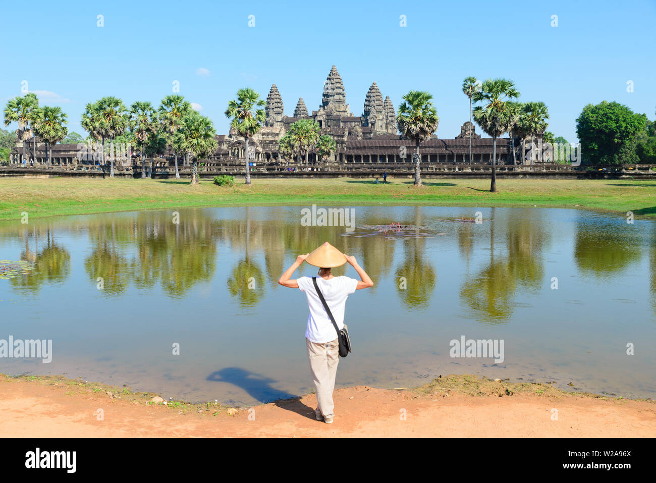 Eine touristische besuchen die Ruinen von Angkor Wat bei Sonnenaufgang, Reiseziel Kambodscha. Frau mit traditionellen Hut und erhobenen Armen, Rückansicht, Hauptfassade refl Stockfoto