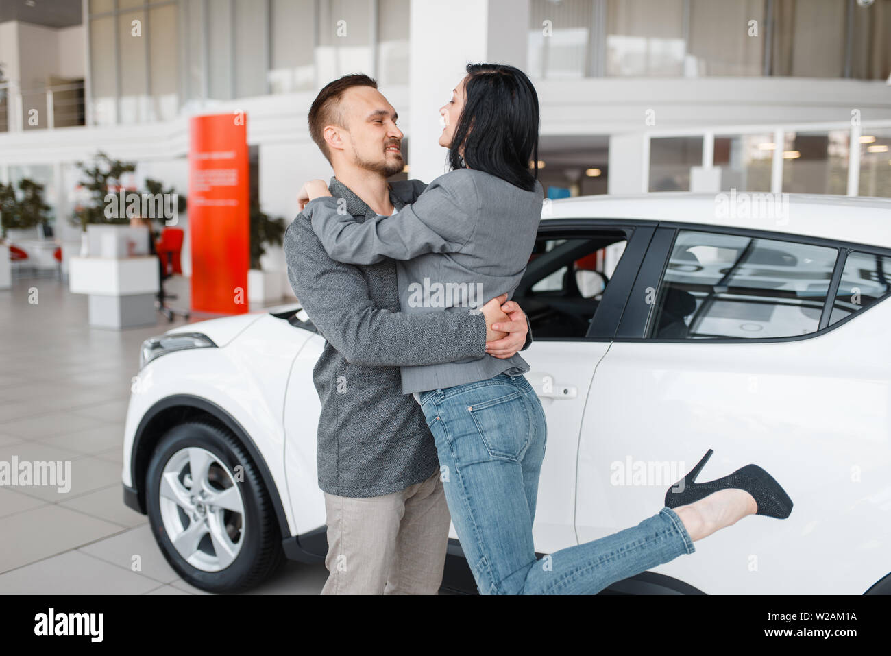 Glückliches Paar Kauf neuer Autos im Showroom, Mann und Frau, umarmen sich. Männliche und weibliche Kunden, Fahrzeug im Autohaus, Auto Verkauf, Auto günstig online Stockfoto